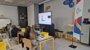 Uczniowie siedzą w ławkach i uczą się programowania z użyciem klocków LEGO SPIKE, w tle jest tablica multimedialna – 