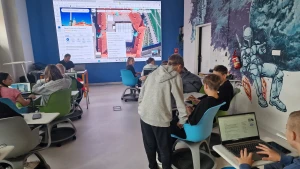 Uczniowie siedzą w ławkach, przed nimi jest ekran z mapami Google – 