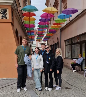 Grupa młodzieży stoi w uliczce z kolorowymi parasolami nad głową –